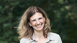 Eine Frau mit schulterlangem, hellbraunem Haar lächelt herzlich in die Kamera. Sie trägt eine helle Bluse und steht im Freien vor einem unscharfen, grünen Waldhintergrund, der eine ruhige und natürliche Atmosphäre schafft.