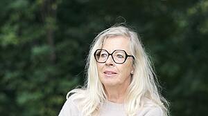 Eine Frau mit langen, hellblonden Haaren und runder Brille trägt einen hellen Pullover. Sie blickt mit freundlichem Gesichtsausdruck in die Kamera. Im Hintergrund ist ein unscharfer, grüner Wald zu sehen.